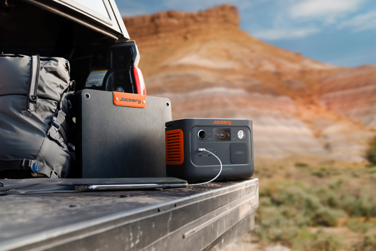 Kleine orange schwarze Jackery Powerstation mit angeschlossenem Smartphone auf offenem Autoheck vor Wüstenkulisse