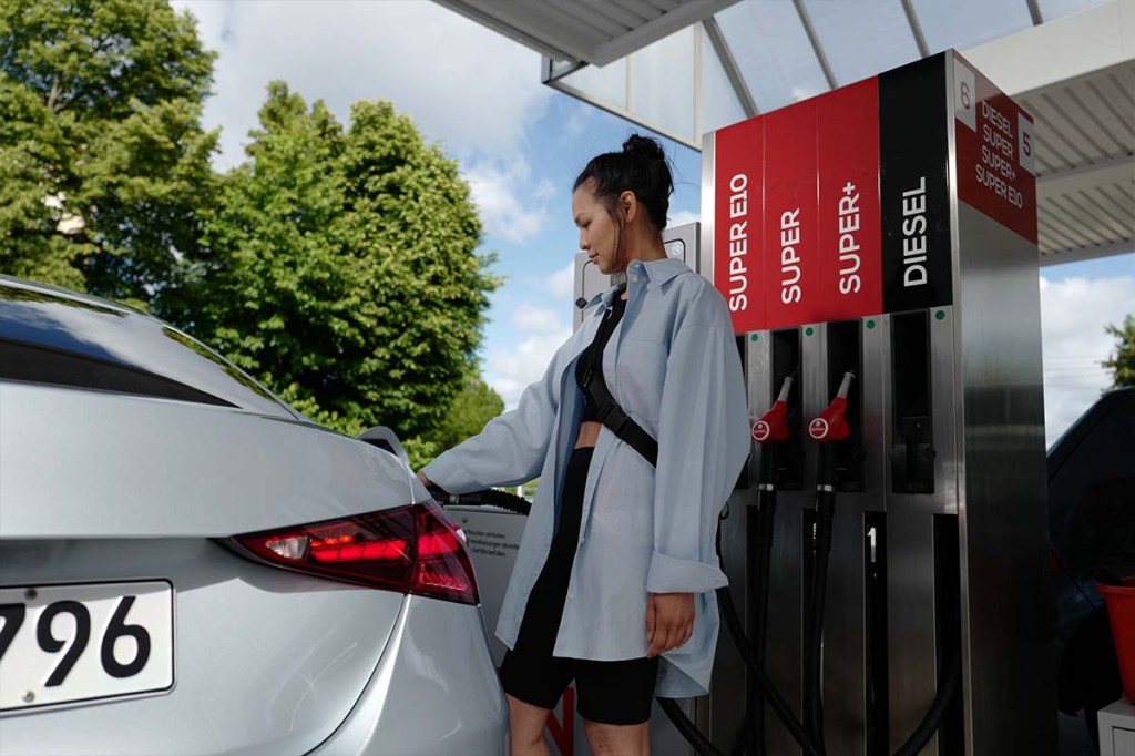 Frau steht neben ihrem Auto, welches gerade an einer Tankstelle betankt wird