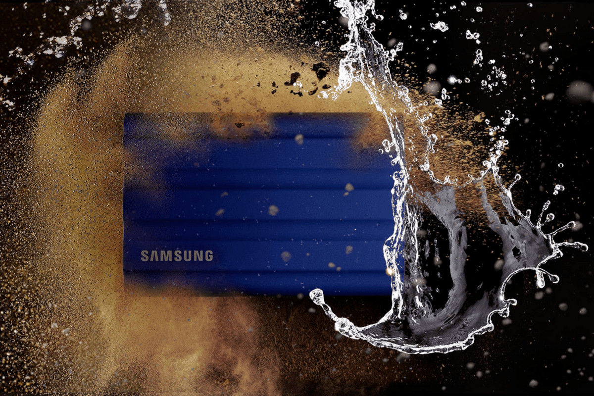 Blaue Samsung T7 Shield rechteckige Festplatte von vorne zwischen Wasser und Sand auf schwarzem Hintergrund