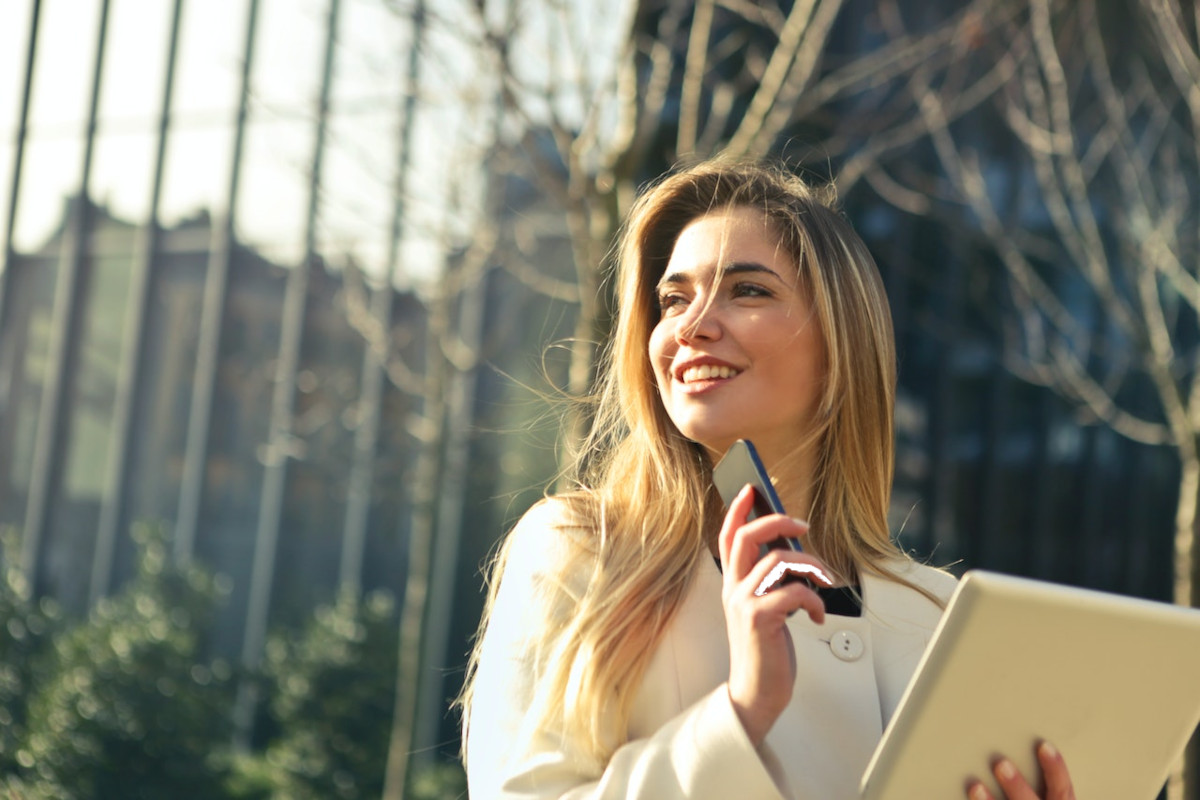 Blonde junge Frau in weißem Pullover hält dunkles Smartphone in der einen und helles Tablet in der anderen Hand und hat den Kopf nach links gedreht und lächelt vor Parkkulisse