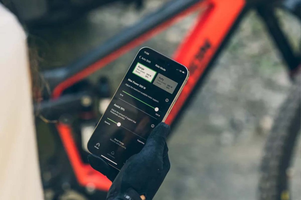 Nahaufnahme Hand hält Smartphone fest, auf dem eine E-Bike App zu sehen ist, im Hintergrund ist ein Fahrrad im Anschnitt zu sehen