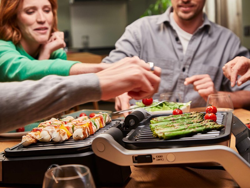 Mehrere Personen sitzen an einem Esstisch, auf dem ein Tefal OptiGrill 4in1 Gemüse und Fleischspieße zubereitet.