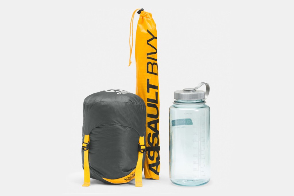 Grau gelber zusammengepackter Biwaksack neben länglichem gelben Beutel und ungefüllter durchsichtiger 1 Liter Wasserflasche