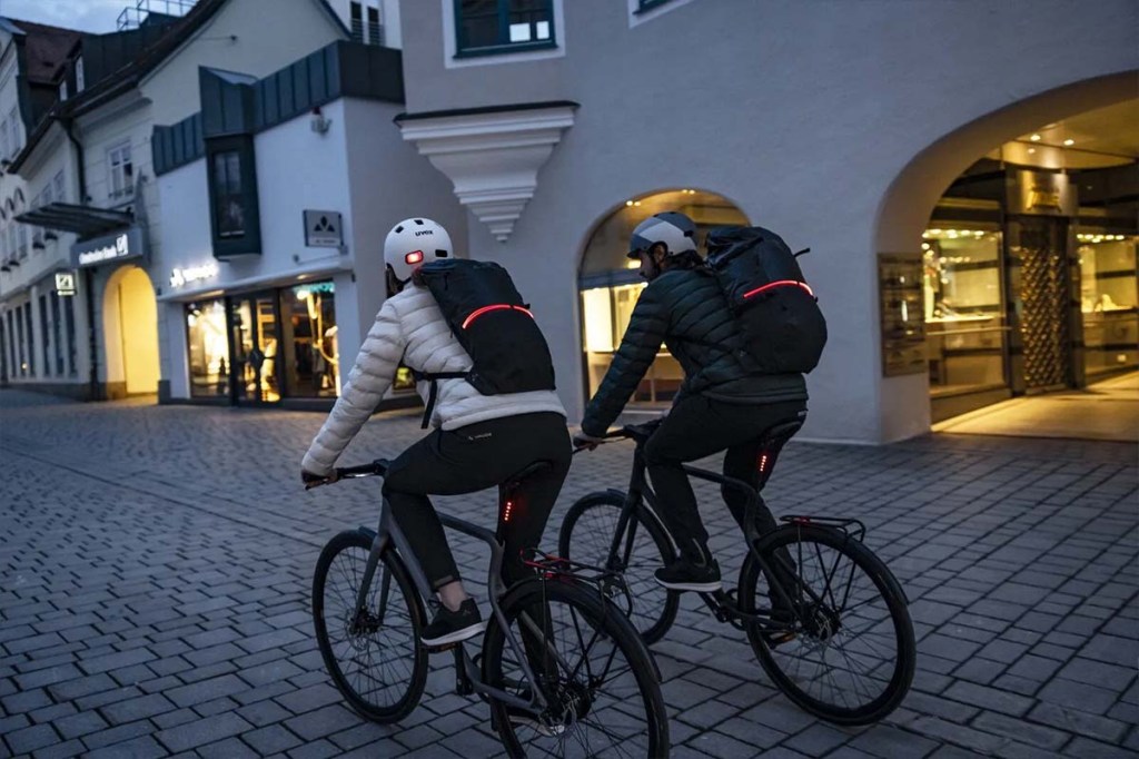 Zwei Personen fahren mit dem Fahrrad durch die Stadt, man sieht sie von hinten, sie haben eine Rucksack auf, der mit einem leuchtenden LED-Band umwickelt ist