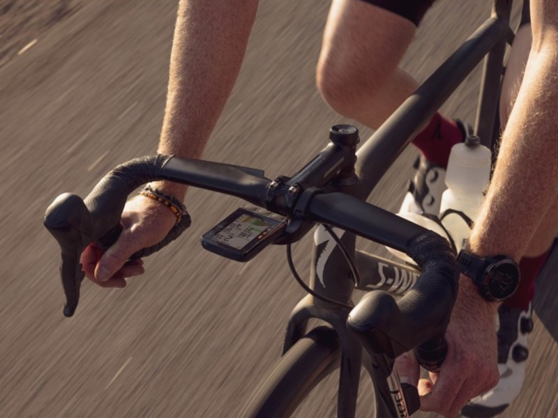 Schwarzes Rennrad fährt auf Asphalt und ragt von rechts oben ins Bild, Lenker wird von zwei Armen gehalten, am Lenker das rechteckige Fahrrad-Navi von Wahoo