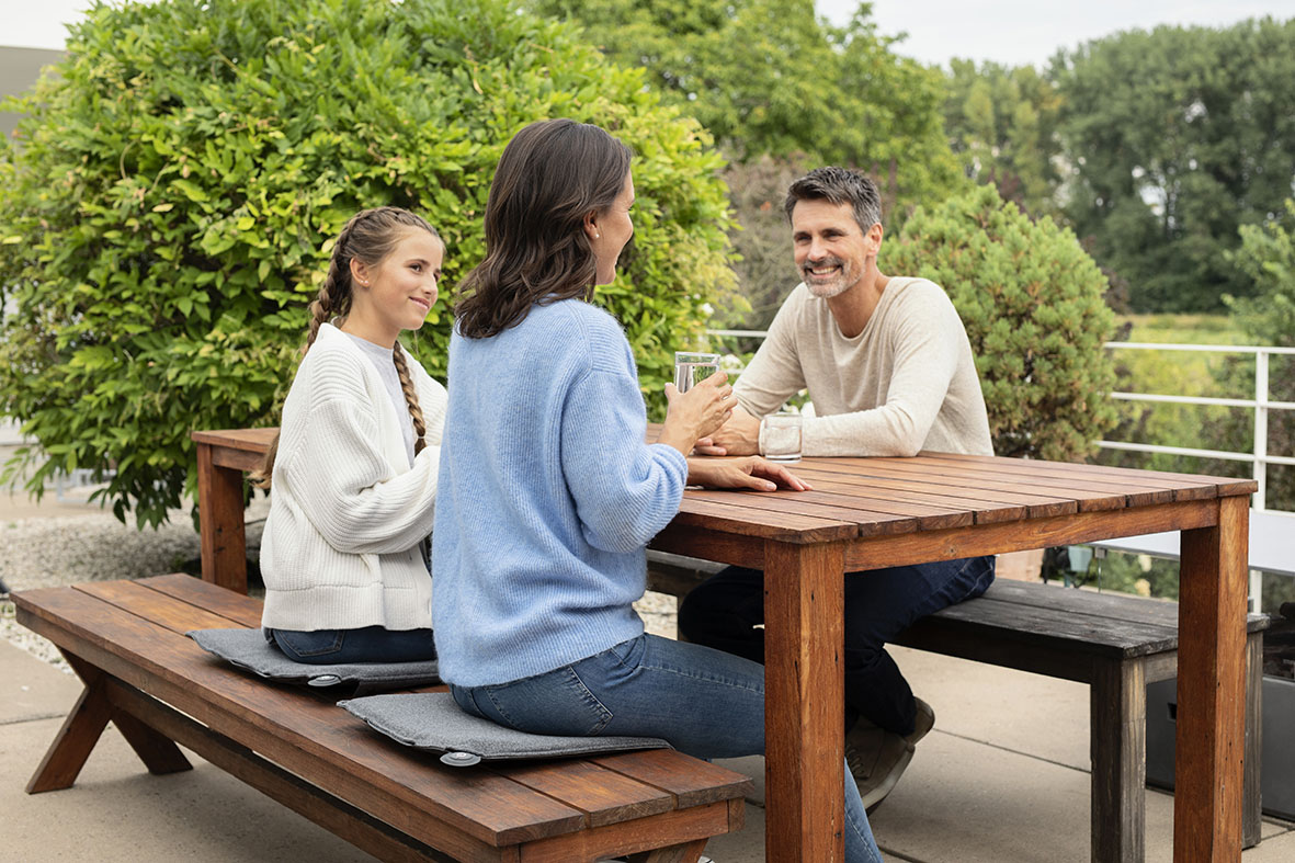 Frau, Mädchen und Mann sitzen auf einer Terrasse an einem Holztisch auf Wäremkissen.