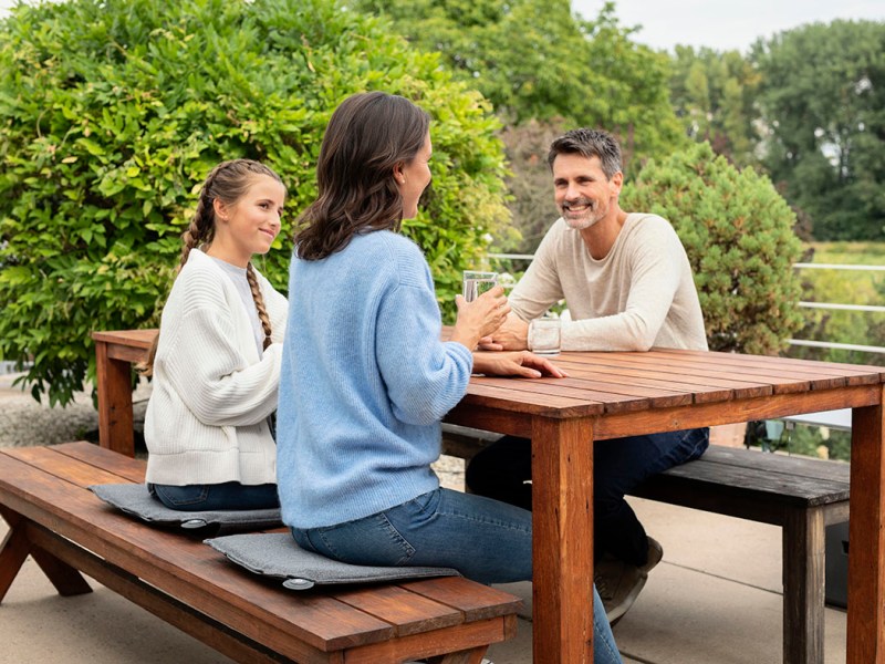 Frau, Mädchen und Mann sitzen auf einer Terrasse an einem Holztisch auf Wäremkissen.