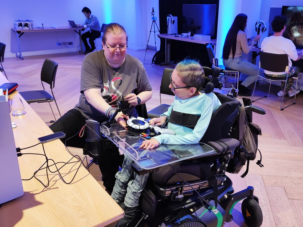 Eine Frau im Rollstuhl spielt mit Hilfe des Access Controllers Sackman. Ihre Assistentin hilft ihr.