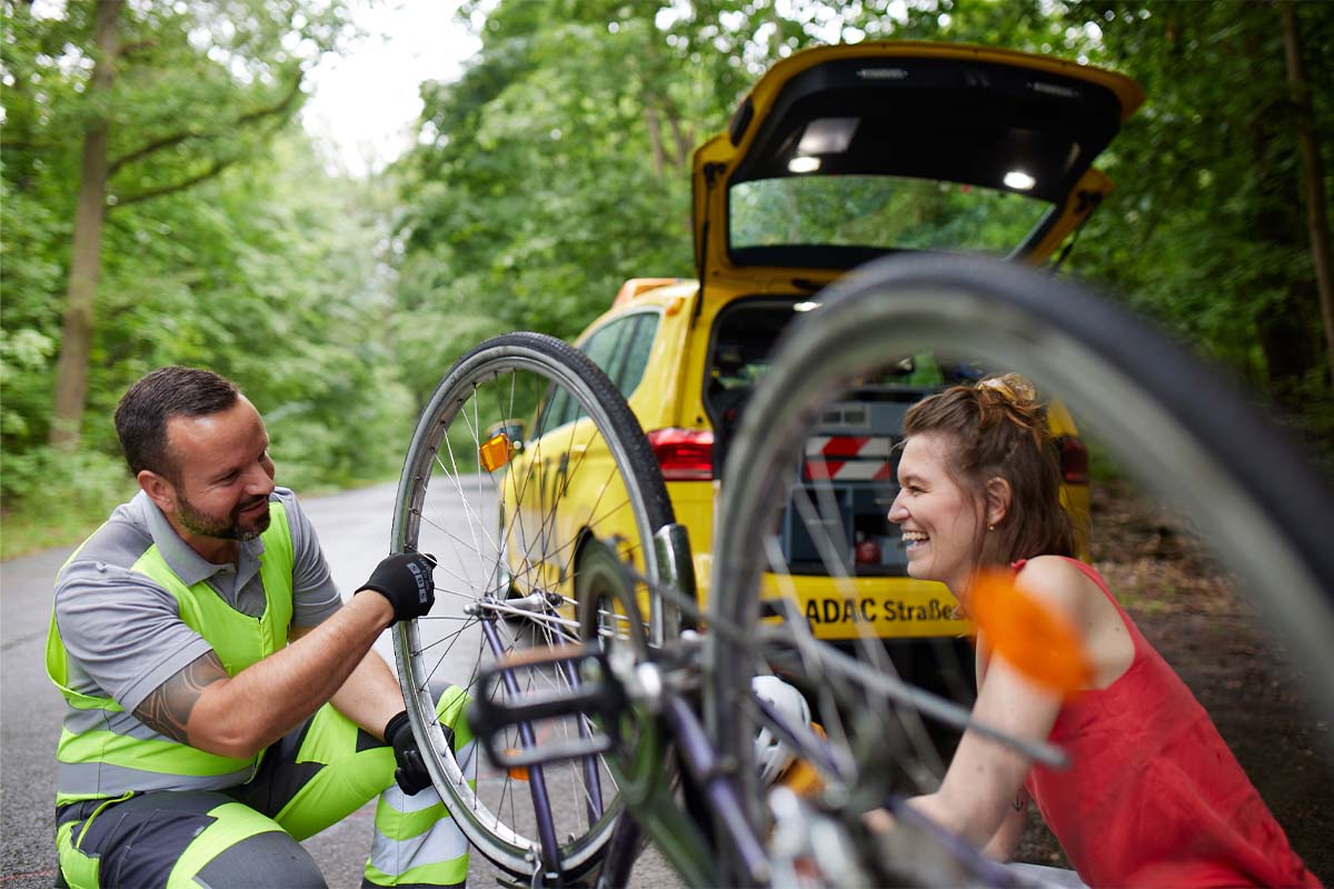 ADAC-Mitarbeiter repariert Fahrrad, lächelnde Frau und ADAC-Auto im Hintergrund