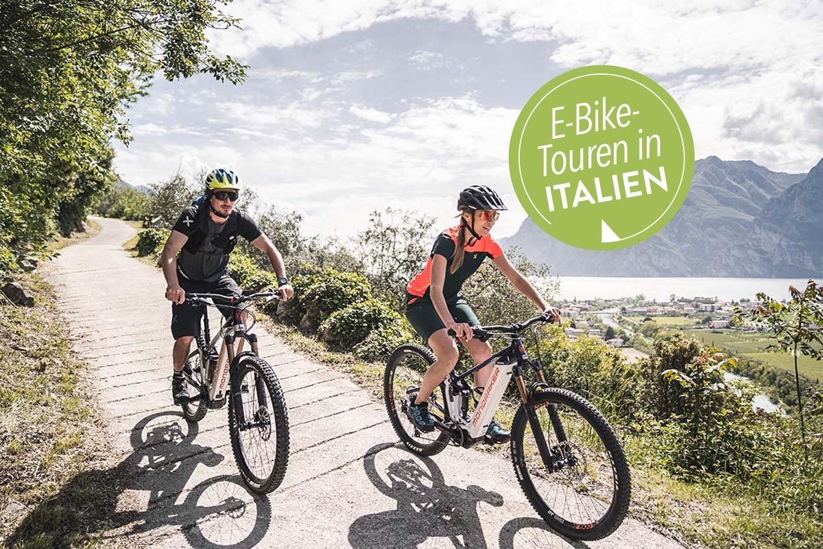 Zwei Fahrradfahrer fahren entlang einer malerischen Naturlandschaft, im Hintergrund ein See und Berge