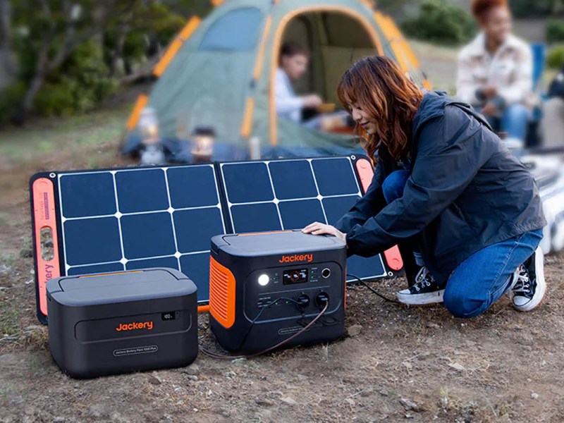 Eine Person schließt ein Solarpanel an die Jackery Explorer 1000 Plus an, die bereits mit einem Zusatz-Akku erweitert ist. Im Hintergrund ist eine Camping-Szene.