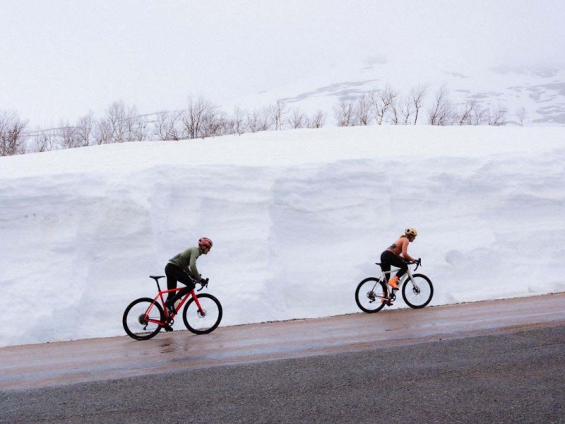 Zwei Radfahrer fahren hintereinander eine Straße lang, hinter ihnen eine Wand aus Schnee