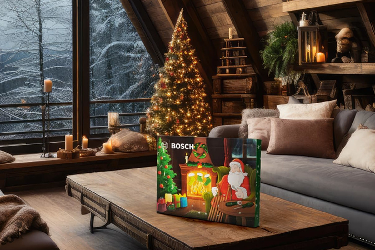 Schräg aufgestellter rechteckiger Adventskalender mit Comic-Weihnachtsmann drauf auf flachem Holztisch, der vor grauem Sofa steht, Holzboden, schräge Holzwand und Fensterfront vor winterlicher Waldkulisse im Hintergrund, daneben ein geschmückter warmleuchtender Tannenbaum