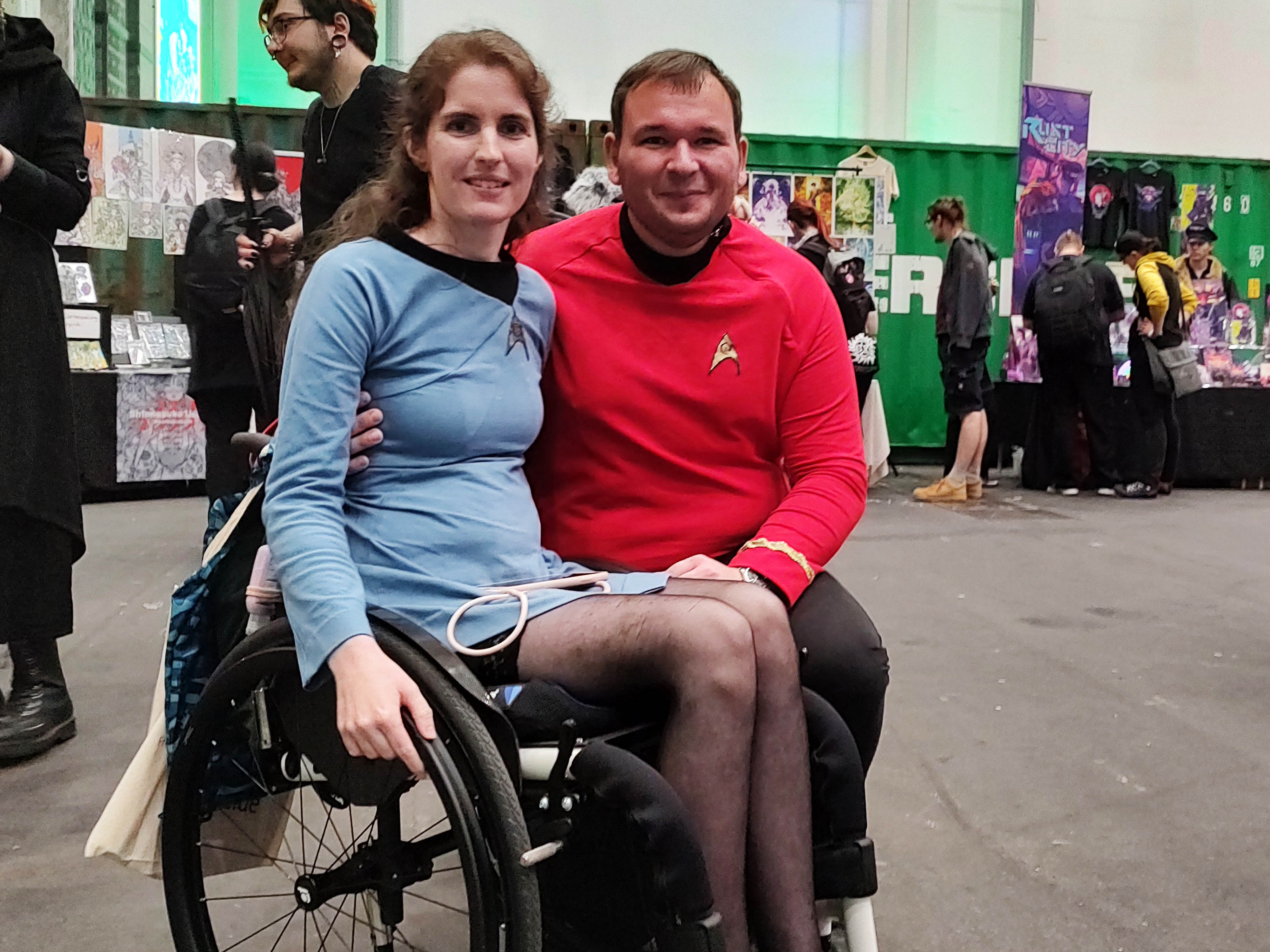 Eine Frau im Rollstuhl und ein Mann tragen Star-Treck-Uniformen in blau und rot.