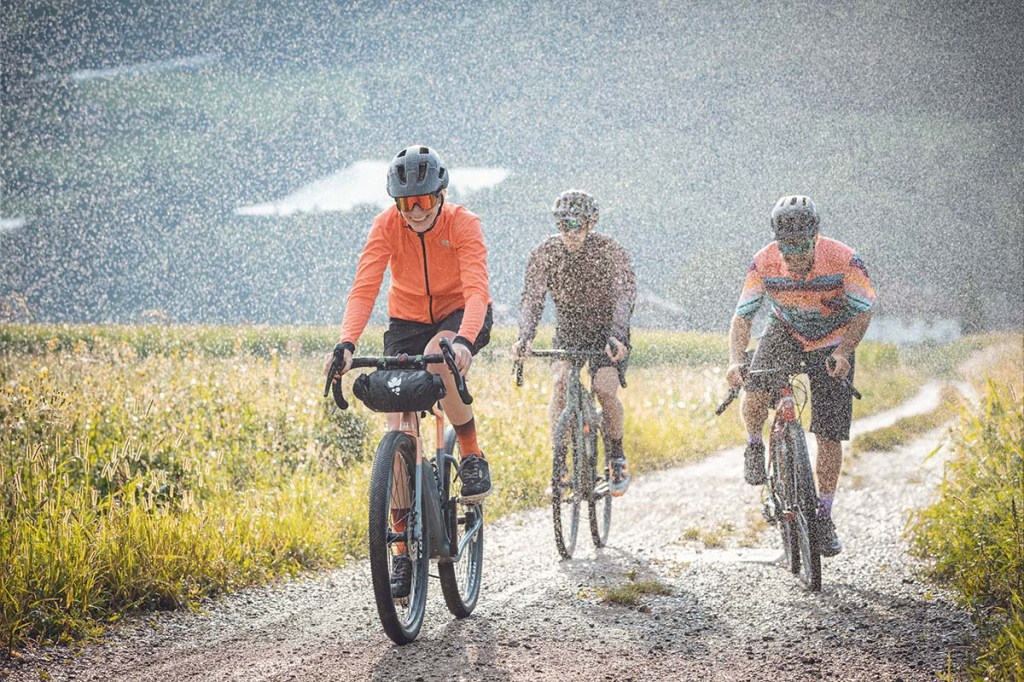 Drei Radfahrer fahren nebeneinander eine Schotterpiste lang, es regnet