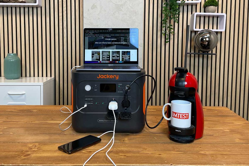 Ein Laptop, ein Smartphone und eine Kaffeemaschine sind an der Jackery-Powerstation angeschlossen, die auf einem Tisch in einer Wohnumgebung steht.