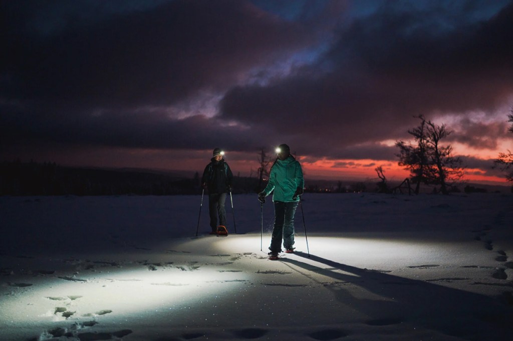 Foto von zwei Personen auf Schneeschuhen mit Skistöcken in den Händen und Stirnlampen auf verschneitem Boden gerichtet. Dunkler Himmel mit Wolken, am Horizont leicht oranges Sonnenlicht