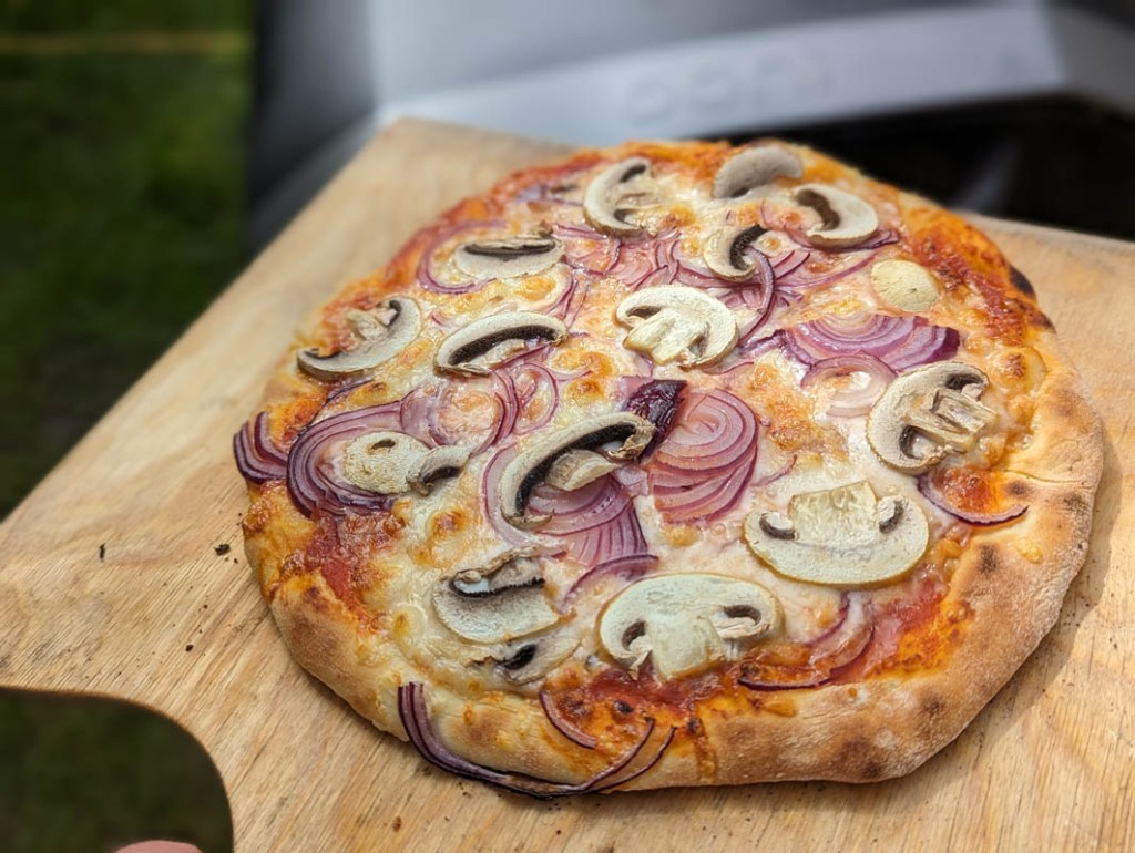 Eine fertige Pizza mit Zwiebeln und Champignons auf einem Holzschieber. Im Hintergrund der Ooni Volt 12.