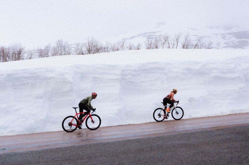 Zwei Radfahrer fahren hintereinander eine Straße entlang, im Hintergrund ist eine Schneemauer