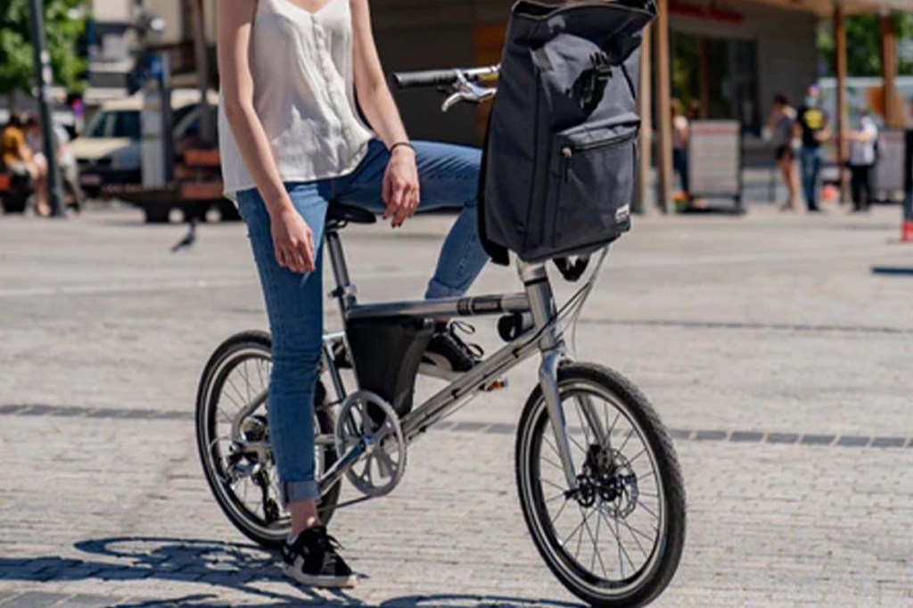 Person, dessen Kopf nicht erkennbar ist, sitzt auf einem Falt-E-Bike, ein Fuß auf dem Boden, einer auf der Pedale