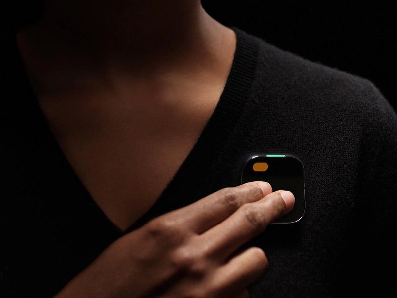 Der Humane Ai Pin, an einem schwarzen Pulli getragen, und mit zwei Fingern berührt.