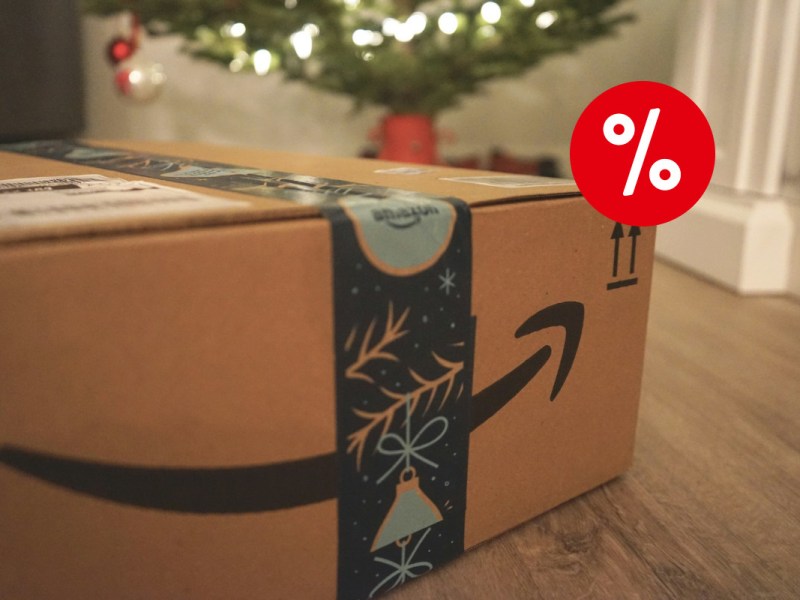 Links unten reinragendes braunes Amazon Paket mit weihnachtlichtem dunklen Paketband auf Holzboden, verschwommen im Hintergrund ein Tannenbaum, mit rotem Prozentzeichen oben rechts