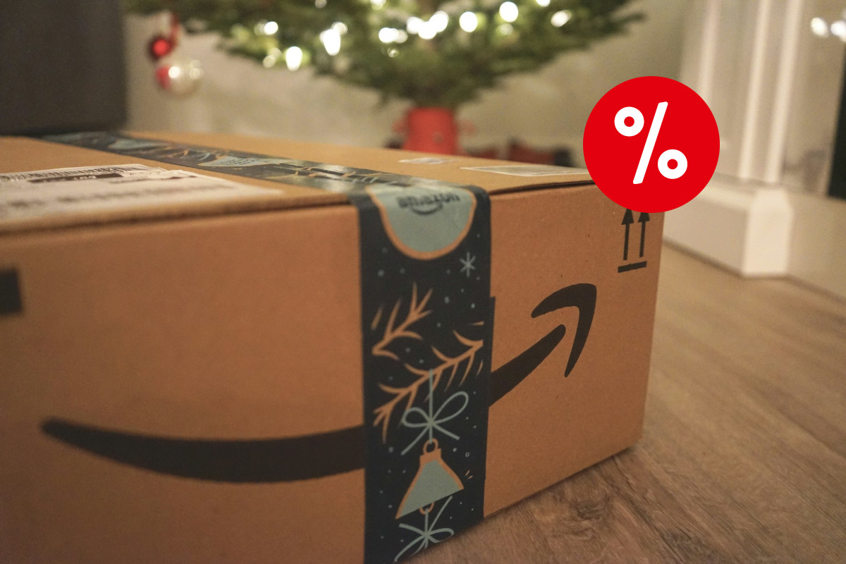 Links unten reinragendes braunes Amazon Paket mit weihnachtlichtem dunklen Paketband auf Holzboden, verschwommen im Hintergrund ein Tannenbaum, mit rotem Prozentzeichen oben rechts