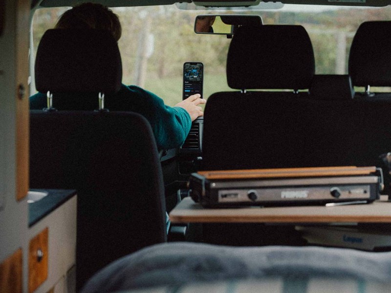 Blick von Heck eines Wohnmobils nach vorne raus, man sieht, dass eine Person mit dem Finger auf ein Smartphone tippt, welches an der Windschutzscheibe befestigt ist