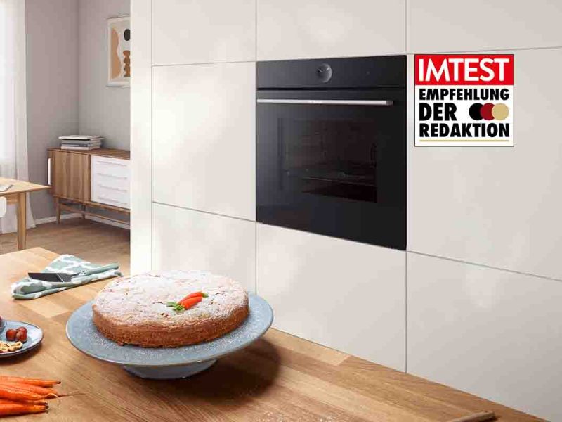 Der Bosch-Einbaubackofen in einer weißen Küchenzeile. Auf der Arbeitsfläche davor stehen ein gebackener Möhrenkuchen sowie Backutensilien. Das IMTEST-Siegel "Empfehlung der Redaktion" ist zu sehen.