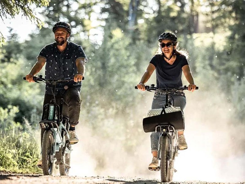 Zwei Menschen fahren mit einem E-Bike nebeneinander durch die Natur, man sieht sie von vorne auf die Kamera zukommen