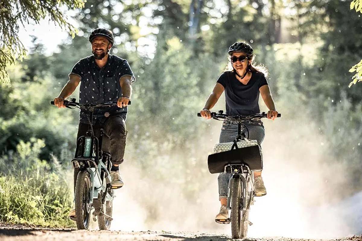 Zwei Menschen fahren mit einem E-Bike nebeneinander durch die Natur, man sieht sie von vorne auf die Kamera zukommen
