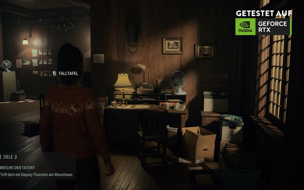 Screenshot aus Alan Wake 2. Eine Person steht an einem Schreibtisch, der Raum ist düster beleuchtet, Licht fällt durch das Fenster rechts.