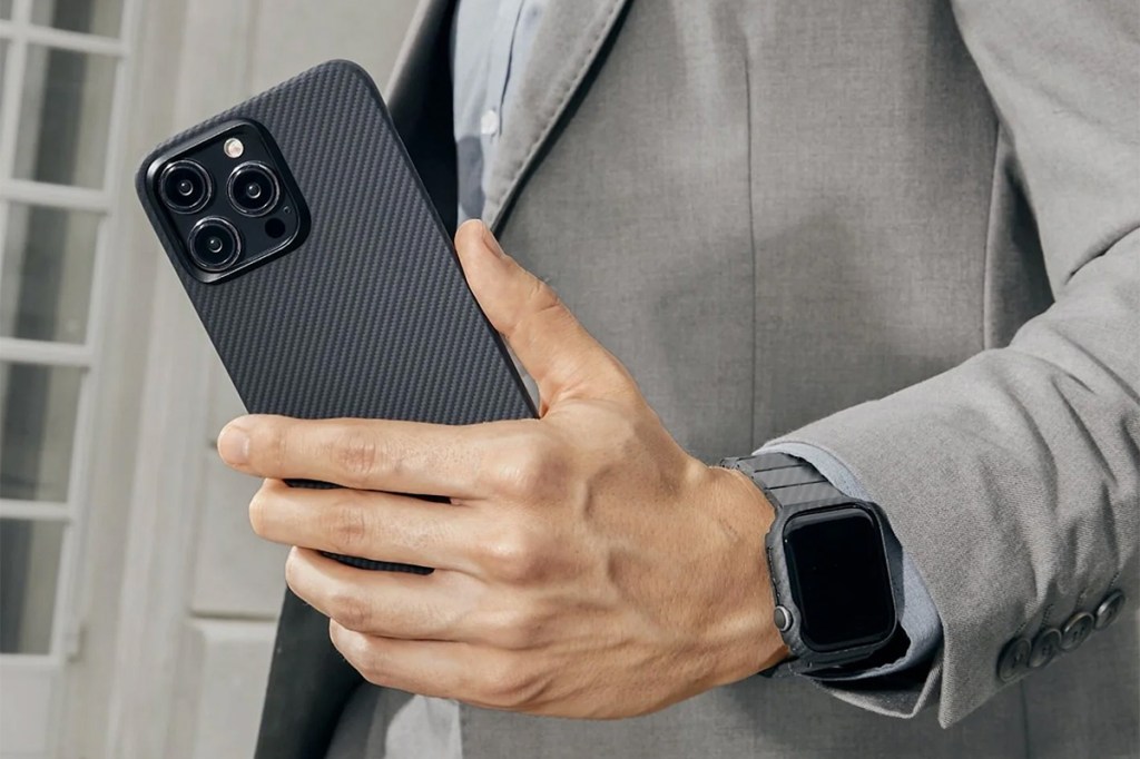 Foto von einer Männerhand mit einem iPhone 15 samt Hülle, außerdem ist eine Smartwatch am Handgelenk zu sehen.