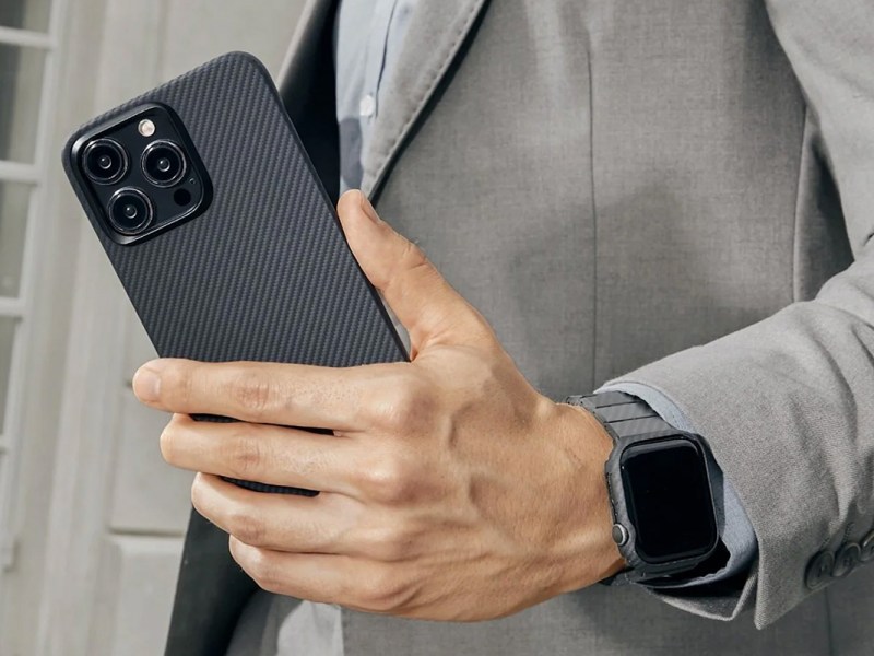 Foto von einer Männerhand mit einem iPhone 15 samt Hülle, außerdem ist eine Smartwatch am Handgelenk zu sehen.