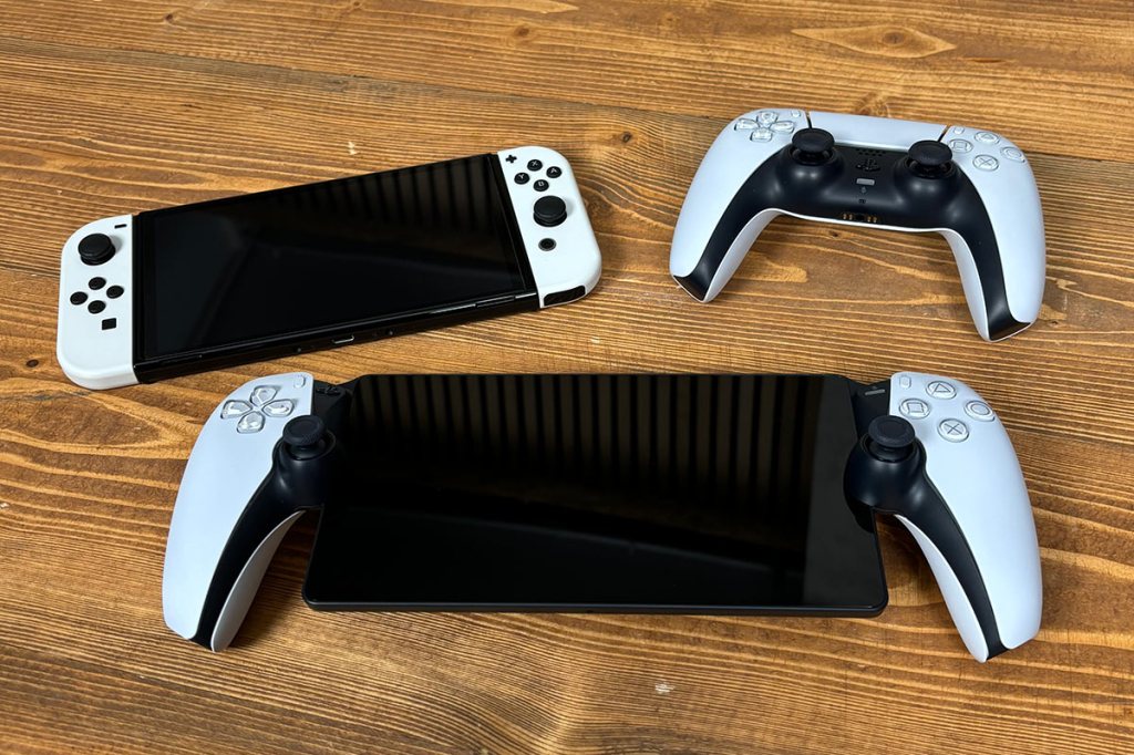 Das PS5-Handheld PlayStation Portal Remote Player, auf einem Holztisch liegend. Daneben liegen, zum Vergleich, die Nintendo Switch OLED und ein PS5-Controller.