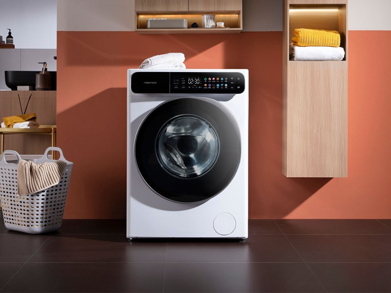 Der Roborock-Waschtrockner auf einem schwarzen Fliesenboden, mit orangener Wand im Hintergrund. Daneben steht ein Wäschekorb.