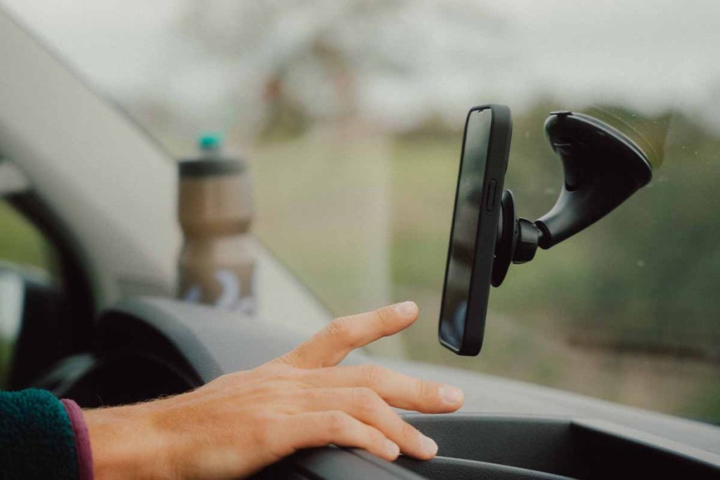 Blick seitlich von innen auf die Windschutzscheibe eines Autos, man sieht eine Smartphonehalterung samt Smartphone an der Windschutzscheibe, eine Finger geht richtung Smartphone