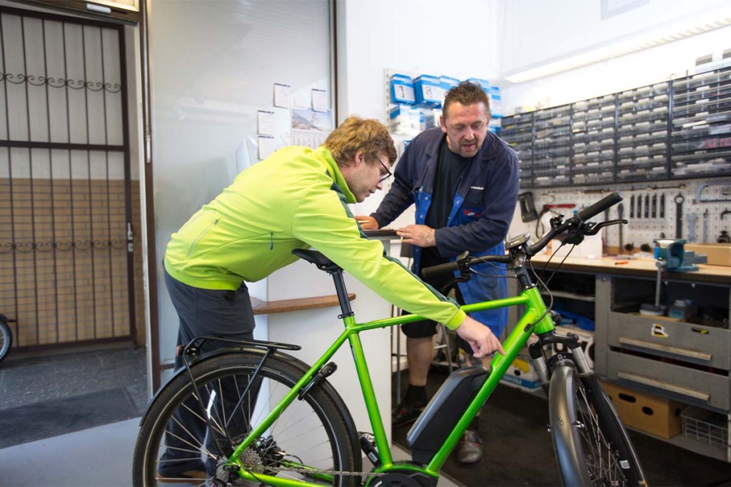 Zwei Männer und ein E-Bike in einer Werkstatt, der eine zeigt auf eine Stelle am Fahrrad