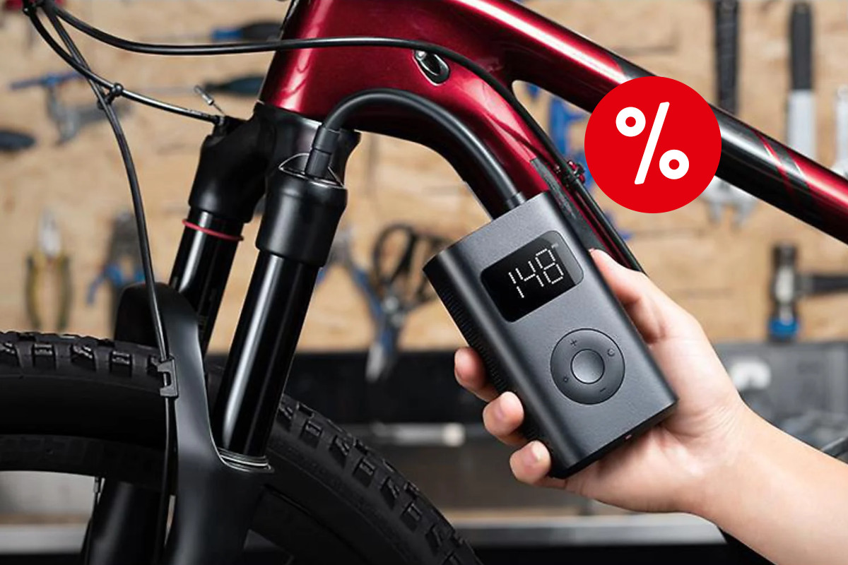Hand rechts im Bild hält schwazren rechteckigen Xiaomi-Kompressor, der an Fahrradreifen von rotem Fahrrad angeschlossen ist; rotes Prozentzeichen rechts oben