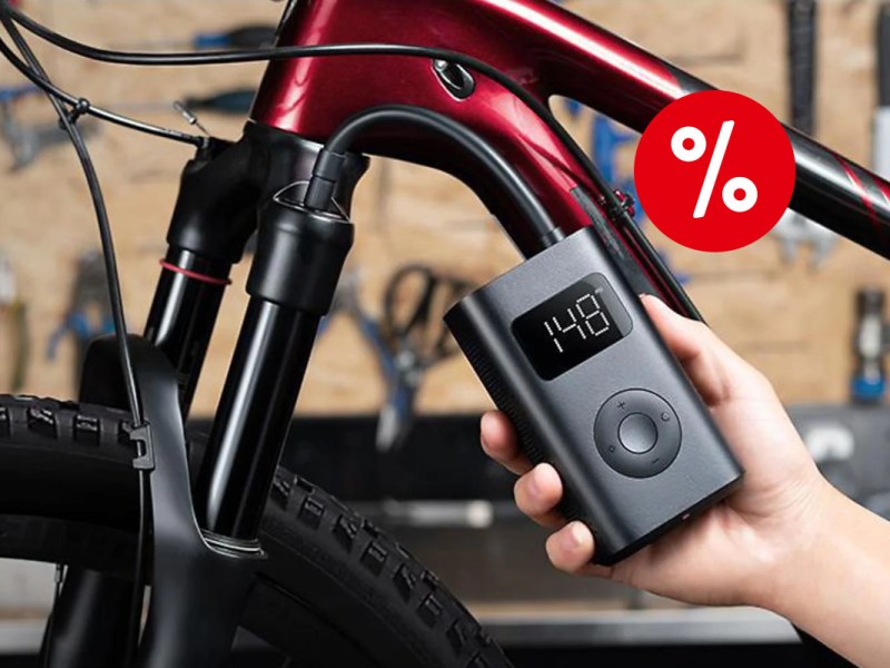 Hand rechts im Bild hält schwazren rechteckigen Xiaomi-Kompressor, der an Fahrradreifen von rotem Fahrrad angeschlossen ist; rotes Prozentzeichen rechts oben