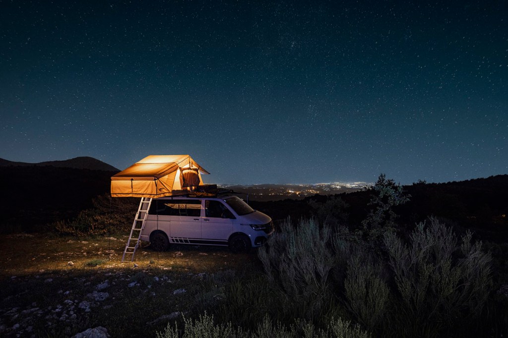 Dachzelt auf VW-Bus bei Nacht mit Beleuchtung vor Berg-Kulisse.