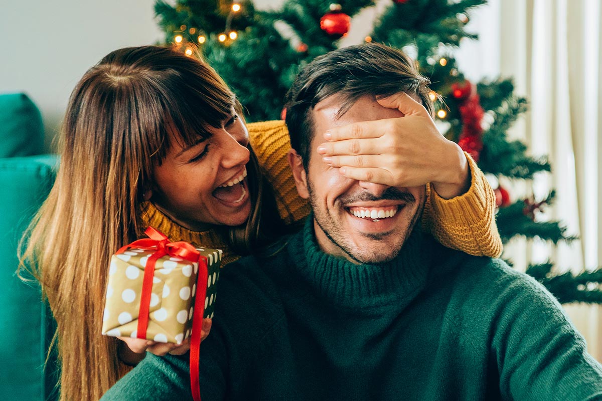 Eine Frau hält einem Mann unter dem Weihnachtsbaum bei der Bescherung lachend die Augen zu.