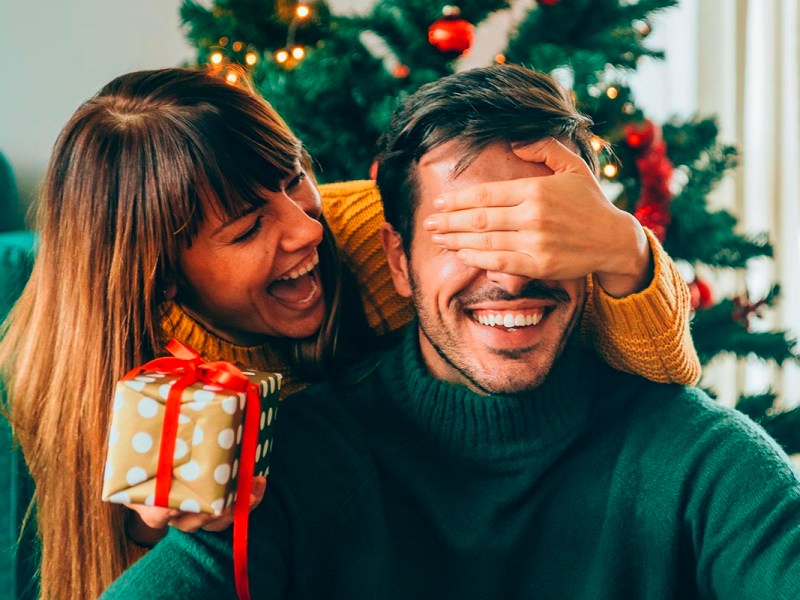 Eine Frau hält einem Mann unter dem Weihnachtsbaum bei der Bescherung lachend die Augen zu.