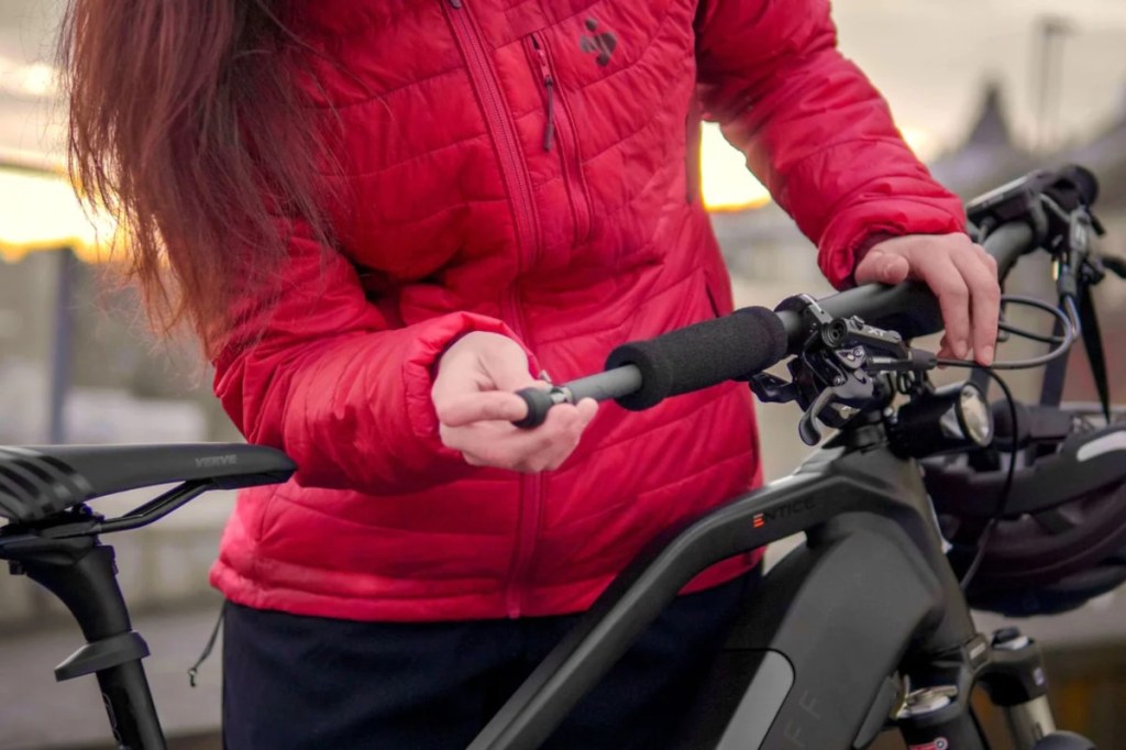 Person in roter Jacke mit roten langen Haaren hinter schwarzem Fahrrad präpariert den Lenker und schiebt grauen länglichen Sender hinein