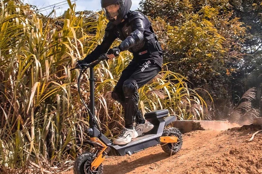 E-Scooter Fahrer mit Scooter auf einer Sandpiste fahrend, dahinter sieht man Sträucher