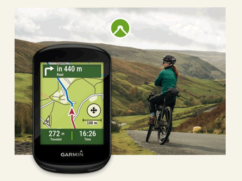 Garmin-Navigationsgerät mit Karte im Display, vor dem HIntergrund einer Radfahrerin in einer Berglandschaft.