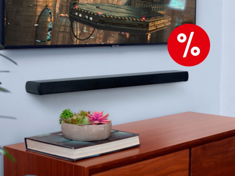 hellblaue Wand mit schwarzer länglicher JBL-Soundbar in der Mitte, darüber ein Fernseher mit Raumschiff-Bild, dadrunter eine Holzkommode mit Buch und Pflanzenschale, rechts oben ein rotes Prozentzeichen