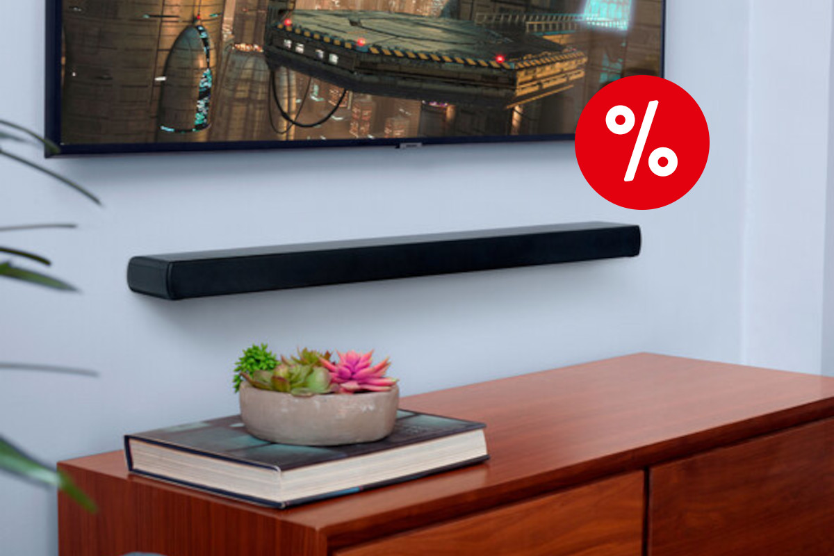 hellblaue Wand mit schwarzer länglicher JBL-Soundbar in der Mitte, darüber ein Fernseher mit Raumschiff-Bild, dadrunter eine Holzkommode mit Buch und Pflanzenschale, rechts oben ein rotes Prozentzeichen