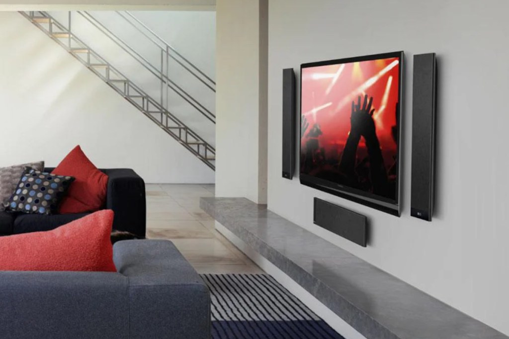 Rechts unten im Bild eine blaue eckige Couch mit roten Kissen vor einem Fernseher, der ein rot schwarzes Bild von Konzert zeigt umrahmt von drei flaachen eckigen Lautsprechern, die zu einem 5.1-Soundsystem gehören, an grauer Wand mit grauem glänzenden Sideboard darunter