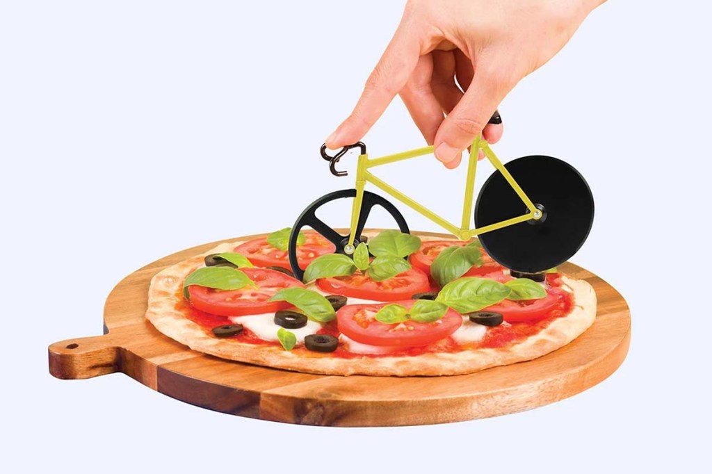 Pizza auf einem Holzbrettchen liegend, ein Pizzaschneider in Form eines Fahrrads wird von einer Hand darüber gefahren.